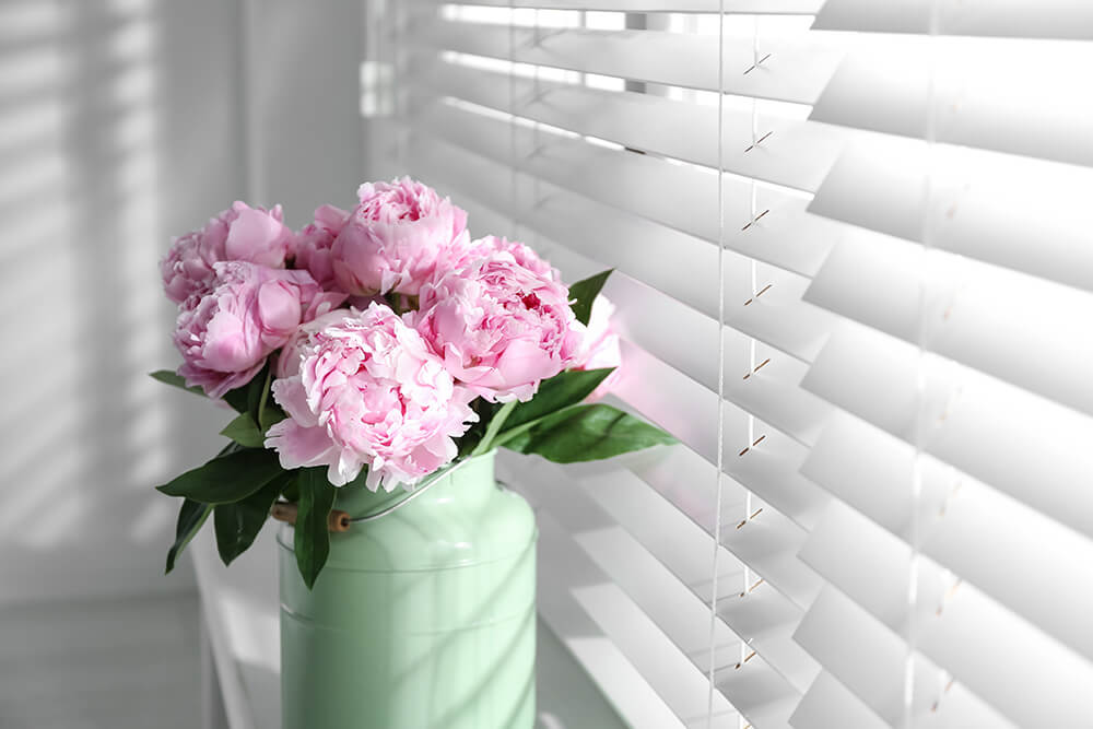 White open blinds with pretty pink flowers on the window sill and light streaming in